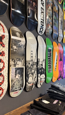 A variety of skateboard decks are displayed vertically on a wall. Each deck features unique designs and graphics, including black and white photography, colorful abstract patterns, and text. The decks are arranged in a row, showcasing their different artistic styles.