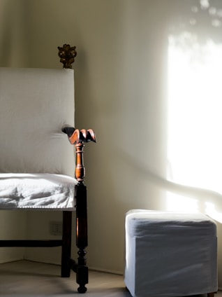 A vintage wooden chair with ornate carvings at the top and armrest stands in a softly lit room. Beside it, a small white ottoman is positioned close to a wall where sunlight filters through, casting gentle shadows.