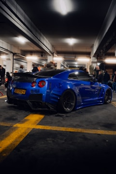 A heavily modified blue sports car is parked in a dimly lit underground garage. Several people are standing around, some looking at the car and others engaged in their own activities. The garage has a concrete floor with yellow lines marking the parking spaces.