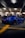 A heavily modified blue sports car is parked in a dimly lit underground garage. Several people are standing around, some looking at the car and others engaged in their own activities. The garage has a concrete floor with yellow lines marking the parking spaces.