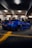 A heavily modified blue sports car is parked in a dimly lit underground garage. Several people are standing around, some looking at the car and others engaged in their own activities. The garage has a concrete floor with yellow lines marking the parking spaces.