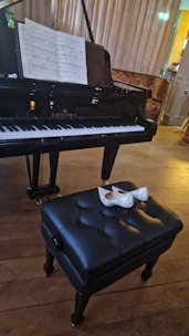 A grand piano with sheet music on the stand, set in a warmly lit room. In front of the piano, a black tufted piano bench is visible with a pair of silver glittery high heels resting on it. The floor is wooden, and the background features a soft curtain.