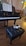A grand piano with sheet music on the stand, set in a warmly lit room. In front of the piano, a black tufted piano bench is visible with a pair of silver glittery high heels resting on it. The floor is wooden, and the background features a soft curtain.