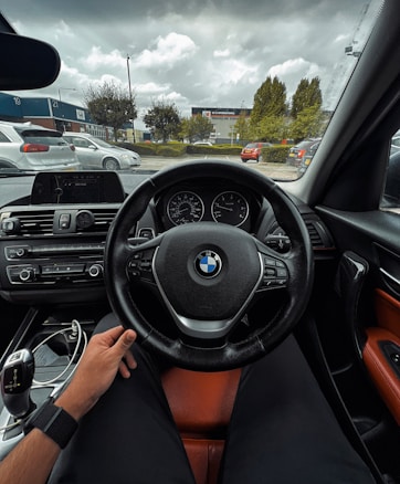 The interior view of a car featuring a steering wheel with the BMW logo, a dashboard with various controls, and a manual gear shift lever. A person's hand is resting on the steering wheel. The car is parked in a lot with visible buildings, trees, and other parked vehicles outside under a cloudy sky.