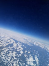 An aerial view displaying a vast landscape covered with a layer of fluffy white clouds beneath an intense blue sky. The curvature of the earth is faintly visible, lending a sense of altitude and expansiveness.