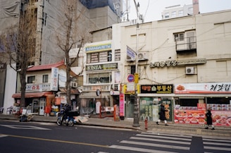 A street scene featuring various storefronts with signs in multiple languages, including Korean and Russian. Buildings are covered with tarps, and trees stand leafless along the sidewalk. A motorcyclist rides along the road while pedestrians walk nearby. The storefronts display a mix of advertisements, with some focusing on travel and dining.