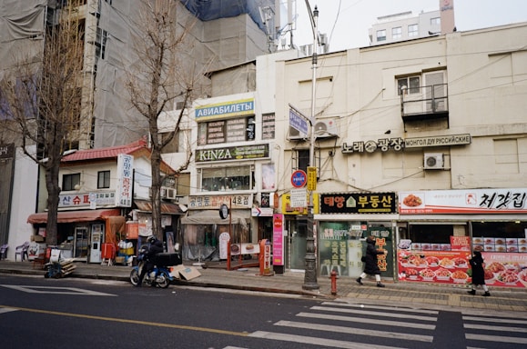A street scene featuring various storefronts with signs in multiple languages, including Korean and Russian. Buildings are covered with tarps, and trees stand leafless along the sidewalk. A motorcyclist rides along the road while pedestrians walk nearby. The storefronts display a mix of advertisements, with some focusing on travel and dining.