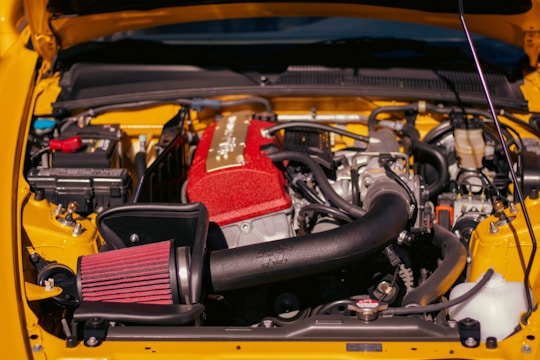 An engine bay of a car with various components visible, including a prominent red air intake filter and a large black air intake tube. The engine cover appears metallic and there are numerous wires and hoses surrounding it. The vehicle's body is painted in a bright yellow color.