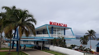 A modern two-story building with blue and white accents situated near a body of water. There is a large bold red sign that reads 'Matanzas' atop the building. The structure is surrounded by a well-maintained yard with palm trees, a manicured lawn, and a white fence. The sky appears overcast, adding a serene atmosphere.