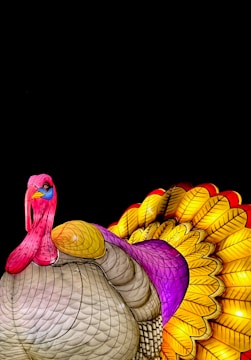 A colorful, artistic depiction of a turkey with its feathers fanned out. The body is predominantly tan with intricate patterns, while the head and neck are painted in bright shades of pink and blue. The tail feathers are vividly detailed, showcasing a gradient of yellow, red, and purple. The dark background enhances the vibrant colors, making the turkey the focal point.