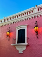 A pink stucco building features a black shuttered window framed with white detailing and a decorative white balcony. On either side of the window, there are two black wrought iron wall lanterns that are lit, giving off a warm orange glow. The sky above is clear and blue.