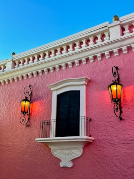 A pink stucco building features a black shuttered window framed with white detailing and a decorative white balcony. On either side of the window, there are two black wrought iron wall lanterns that are lit, giving off a warm orange glow. The sky above is clear and blue.