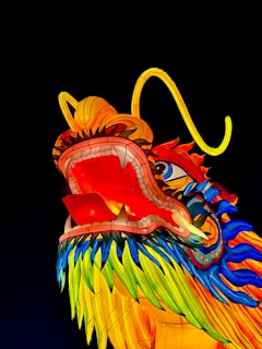 A vibrant and colorful dragon, intricately detailed with bright shades of yellow, red, blue, and green against a dark background. The dragon's mouth is open, displaying sharp teeth and giving it a fierce expression. Its large scaled body is adorned with glowing patterns, and its mane is depicted with elaborate, fiery colors.
