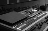 A close-up of a professional audio mixing console with multiple knobs, sliders, and buttons. The image is in black and white and captures the detailed layout of the audio equipment commonly used for sound engineering in a theater or concert setting. In the background, there are blurred rows of seats, indicating a venue environment.