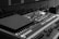 A close-up of a professional audio mixing console with multiple knobs, sliders, and buttons. The image is in black and white and captures the detailed layout of the audio equipment commonly used for sound engineering in a theater or concert setting. In the background, there are blurred rows of seats, indicating a venue environment.