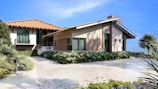 A modern house with a red-tiled roof and beige walls is situated in a landscaped setting. The driveway features neatly trimmed greenery and a parked white car. The blue sky and distant mountains create an inviting and serene ambiance.