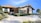 A modern house with a red-tiled roof and beige walls is situated in a landscaped setting. The driveway features neatly trimmed greenery and a parked white car. The blue sky and distant mountains create an inviting and serene ambiance.
