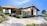 A modern house with a red-tiled roof and beige walls is situated in a landscaped setting. The driveway features neatly trimmed greenery and a parked white car. The blue sky and distant mountains create an inviting and serene ambiance.