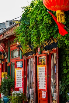 A vibrant street scene with a traditional Asian storefront adorned with red and gold signage. Lush green vines cover part of the building, and a red lantern hangs prominently. The storefront displays intricate decorations and multiple signs written in Chinese characters.