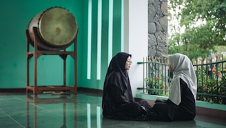 Two women in dark clothing sit facing each other on a reflective floor next to a green wall with vertical white strips. Behind them, there is a large drum on a wooden stand and a railing with greenery outside.