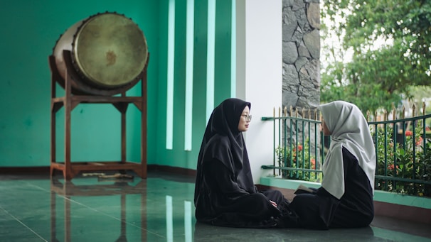 Two women in dark clothing sit facing each other on a reflective floor next to a green wall with vertical white strips. Behind them, there is a large drum on a wooden stand and a railing with greenery outside.