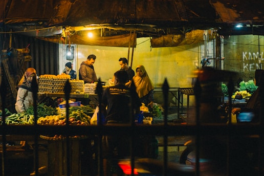 A bustling night market scene with several people interacting around stalls filled with a variety of fresh produce including leafy greens, eggs, and other vegetables. The market is dimly lit with a few overhead lights casting a warm glow over the area. The setting feels intimate and lively with a mix of vendors and customers engaged in transactions.