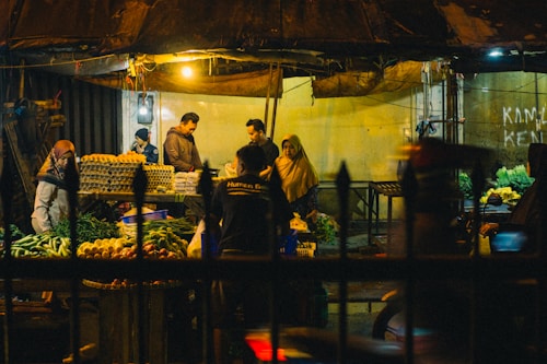 A bustling night market scene with several people interacting around stalls filled with a variety of fresh produce including leafy greens, eggs, and other vegetables. The market is dimly lit with a few overhead lights casting a warm glow over the area. The setting feels intimate and lively with a mix of vendors and customers engaged in transactions.