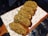 A row of freshly baked bread rolls topped with a crumbly mixture of nuts and herbs is arranged on a wooden serving board lined with decorative paper featuring food-related illustrations.