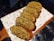 A row of freshly baked bread rolls topped with a crumbly mixture of nuts and herbs is arranged on a wooden serving board lined with decorative paper featuring food-related illustrations.