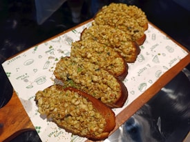 A row of freshly baked bread rolls topped with a crumbly mixture of nuts and herbs is arranged on a wooden serving board lined with decorative paper featuring food-related illustrations.