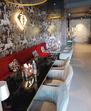 A stylish and modern interior with a long black table surrounded by gray upholstered chairs. The walls feature intricate, whimsical wallpaper with images of unicorns and foliage. A plush red bench adds a pop of color, and above it, a glowing golden logo is mounted on the wall. The space is well-lit, with pendant lights hanging from the ceiling, creating a warm and inviting atmosphere.