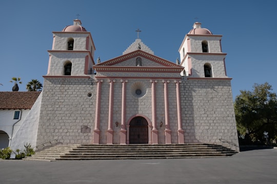 A large, historic church building featuring two bell towers and an ornate facade. The structure is made of stone with pink accents highlighting the architectural details. There are stone steps leading up to a grand wooden door with a circular window above it. The sky is clear, and a few palm trees are visible to the left and right of the church.