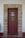 A solid wooden door painted brown is framed by a stone archway with evenly spaced stones. The door features a metal grille covering a small window in the upper half and a simple metal handle on the right side. The surrounding wall is painted white, with a red baseboard near the bottom.