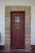 A solid wooden door painted brown is framed by a stone archway with evenly spaced stones. The door features a metal grille covering a small window in the upper half and a simple metal handle on the right side. The surrounding wall is painted white, with a red baseboard near the bottom.