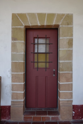 A solid wooden door painted brown is framed by a stone archway with evenly spaced stones. The door features a metal grille covering a small window in the upper half and a simple metal handle on the right side. The surrounding wall is painted white, with a red baseboard near the bottom.