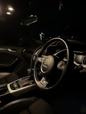 A car interior featuring a modern and sleek design is illuminated by ambient light. The focus is on a steering wheel with a distinctive logo in the center. The dashboard is equipped with various controls and a digital display. The interior is mostly dark with leather seating visible.