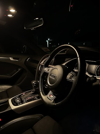 A car interior featuring a modern and sleek design is illuminated by ambient light. The focus is on a steering wheel with a distinctive logo in the center. The dashboard is equipped with various controls and a digital display. The interior is mostly dark with leather seating visible.