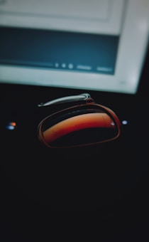 A pair of reflective sunglasses rests on a dark surface, with a computer screen visible in the background. The sunglasses capture a warm reddish reflection, contrasting with the cool tones of the computer screen.