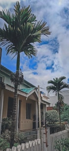 A residential house with a white picket fence and palm trees, set against a partly cloudy blue sky. The house has a beige exterior with a green roof and a front porch. Lush green plants and shrubs are visible near the fence, adding to the tropical ambiance.