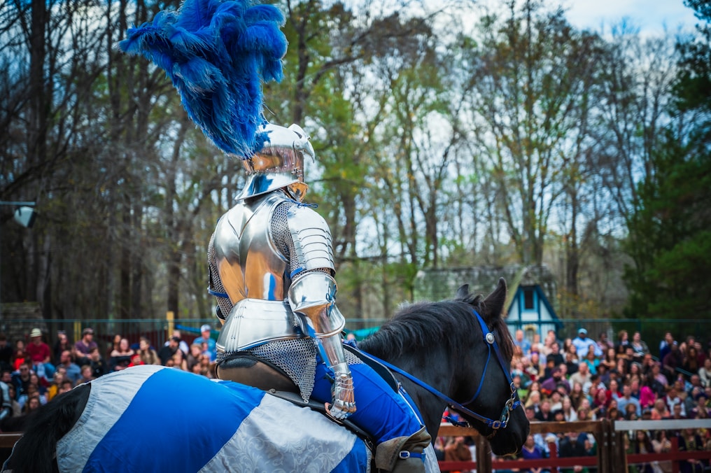 A knight in shining armor with a large blue feathered plume on the helmet is mounted on a horse. The horse is covered with a blue and white caparison. The scene is set outdoors with a wooded background, and there is a large crowd of spectators in the distance, indicating a public event or festival.