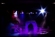 A dimly lit stage with vibrant lighting effects, including spotlights in shades of blue, purple, and white. The central feature is a stone-like archway surrounded by layered platforms. Smoke or mist creates a mystical atmosphere.