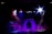 A dimly lit stage with vibrant lighting effects, including spotlights in shades of blue, purple, and white. The central feature is a stone-like archway surrounded by layered platforms. Smoke or mist creates a mystical atmosphere.