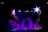 A dimly lit stage with vibrant lighting effects, including spotlights in shades of blue, purple, and white. The central feature is a stone-like archway surrounded by layered platforms. Smoke or mist creates a mystical atmosphere.