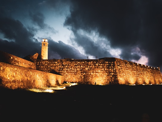 A tall stone wall is highlighted by warm lights against a dramatic evening sky filled with dark, heavy clouds. A clock tower extends from the wall, its face illuminated as dusk settles in. A lone silhouette of a person appears at the top of the wall, adding to the scene's solitary feel.