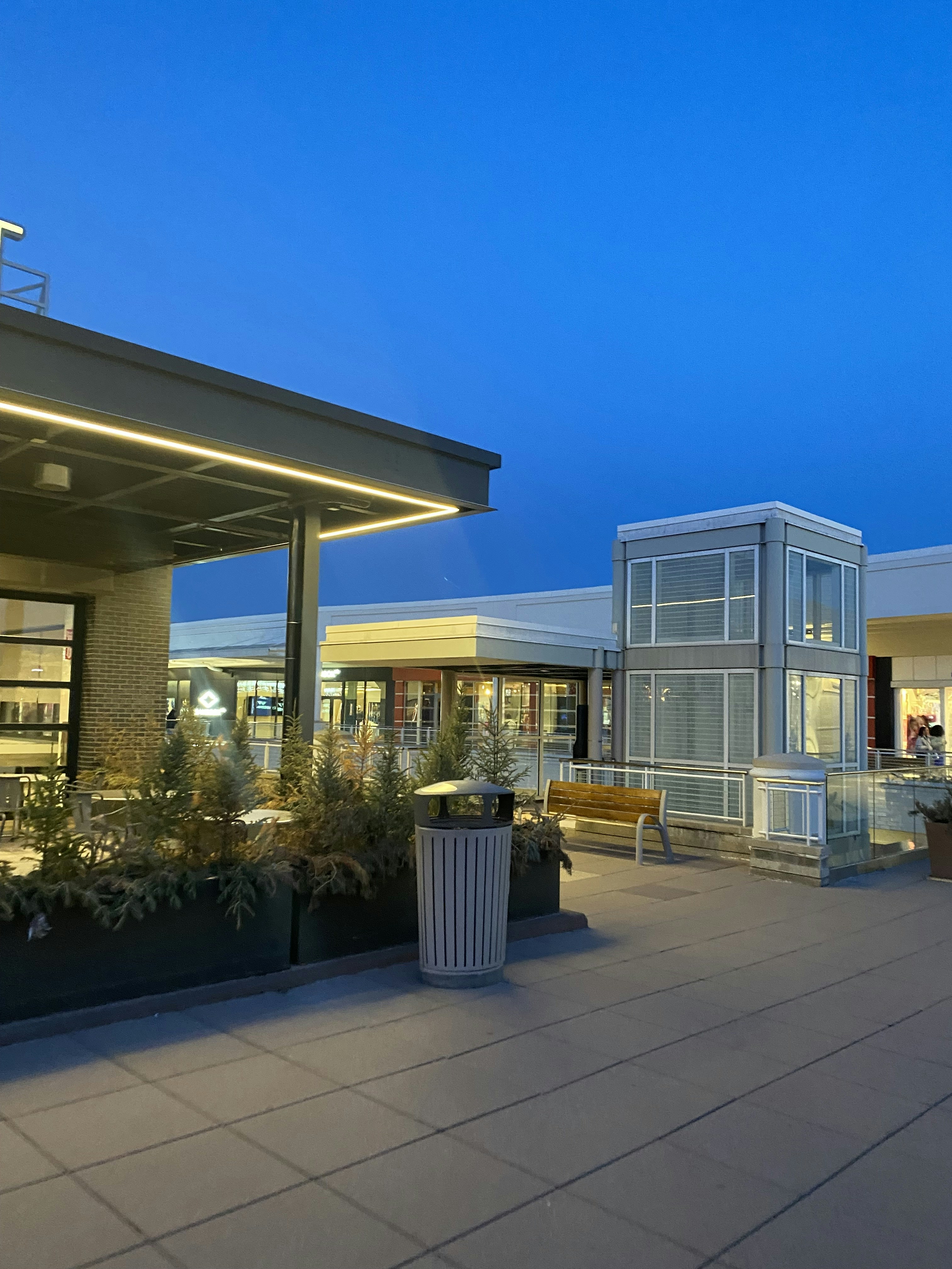 Modern outdoor shopping plaza at dusk, warm ambient lighting, dark building facades, minimal signage