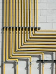 A series of yellow pipes run vertically and horizontally against a light gray concrete block wall, creating a grid-like pattern. The pipes are evenly spaced and neatly arranged, showcasing a structured and industrial design.