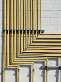 A series of yellow pipes run vertically and horizontally against a light gray concrete block wall, creating a grid-like pattern. The pipes are evenly spaced and neatly arranged, showcasing a structured and industrial design.