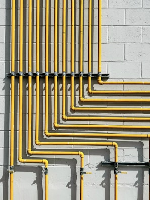 A series of yellow pipes run vertically and horizontally against a light gray concrete block wall, creating a grid-like pattern. The pipes are evenly spaced and neatly arranged, showcasing a structured and industrial design.