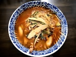 A bowl of spicy soup is filled with a rich, red broth and topped with an assortment of vegetables including mushrooms, cabbage, and green onions. The soup is served in a beautifully patterned blue and white ceramic bowl.