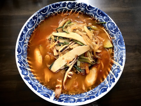 A bowl of spicy soup is filled with a rich, red broth and topped with an assortment of vegetables including mushrooms, cabbage, and green onions. The soup is served in a beautifully patterned blue and white ceramic bowl.